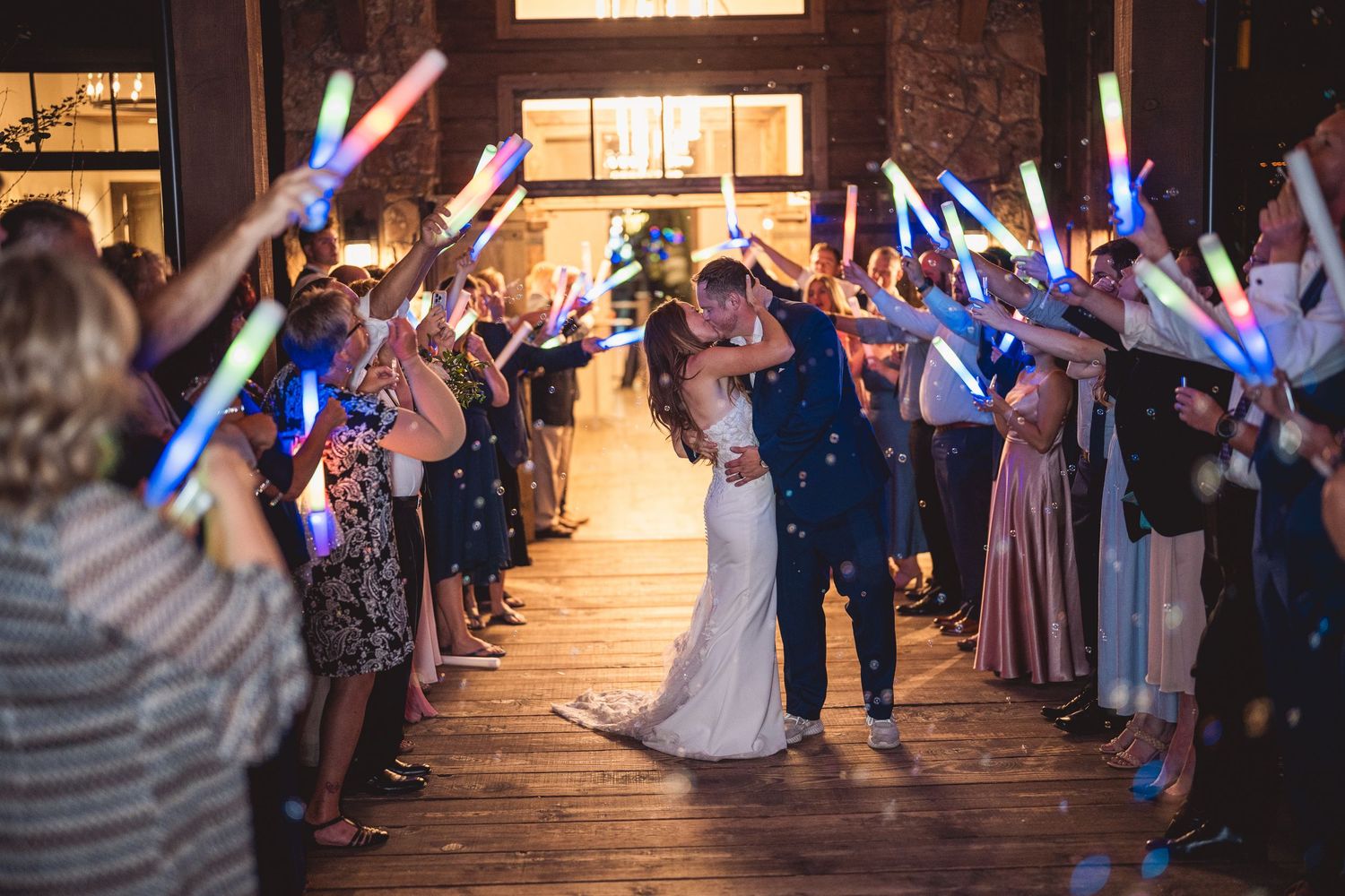 Bride and groom share a kiss during a glowstick send-off at their wedding reception with guests waving colorful LED wands.