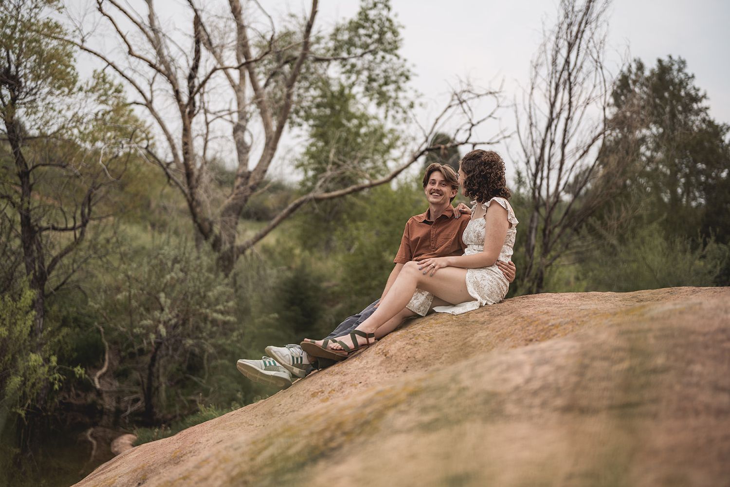 Couple sitting together on a large rock surrounded by lush green trees in a scenic outdoor setting.