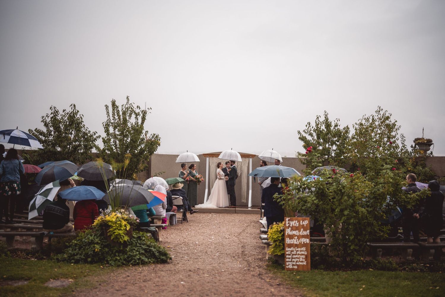 Outdoor wedding ceremony in the rain with guests holding colorful umbrellas, bride and groom at the altar.