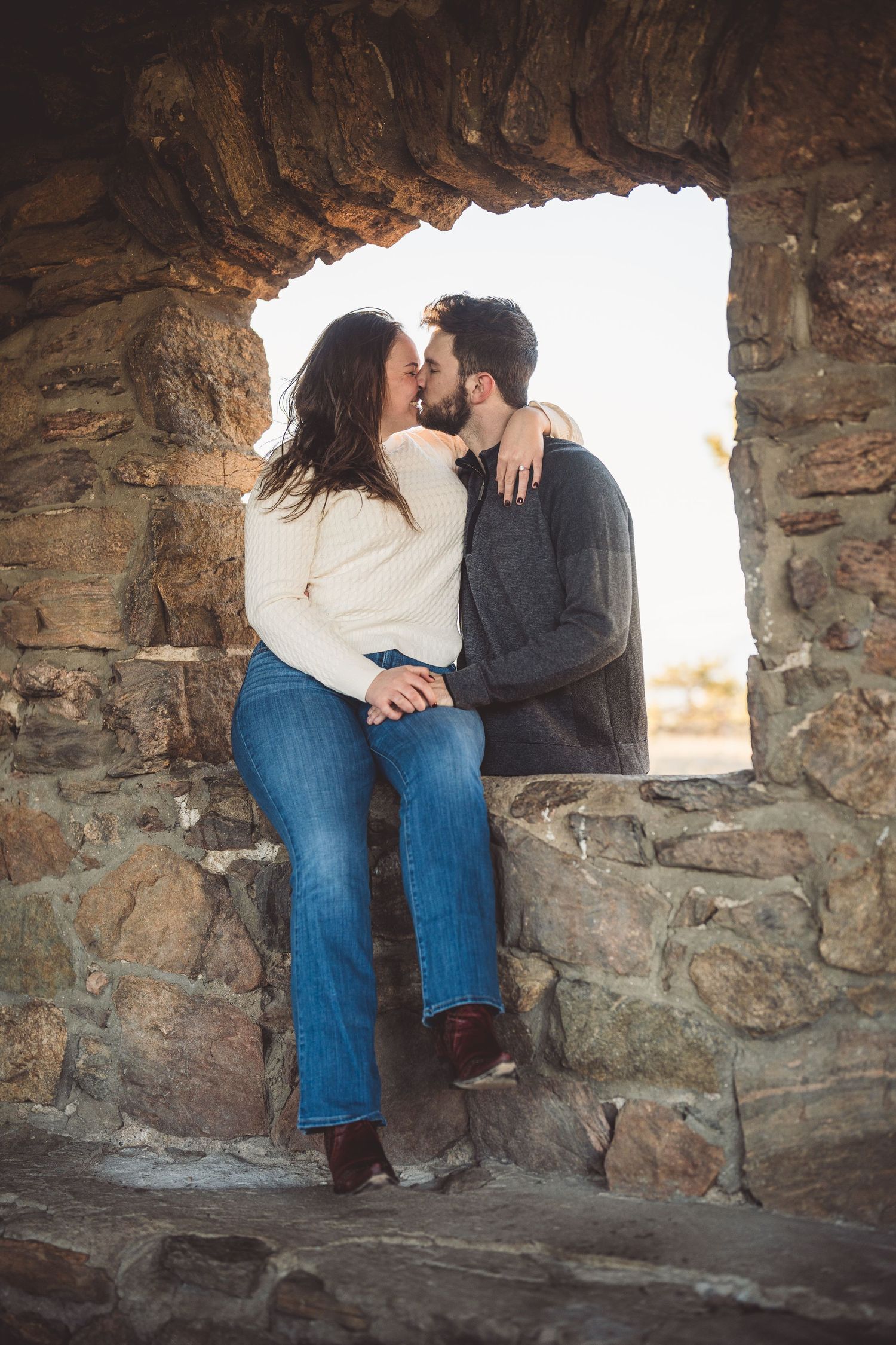 Couple kissing romantically in a rustic stone window arch, woman in white sweater and jeans, man in gray sweater.