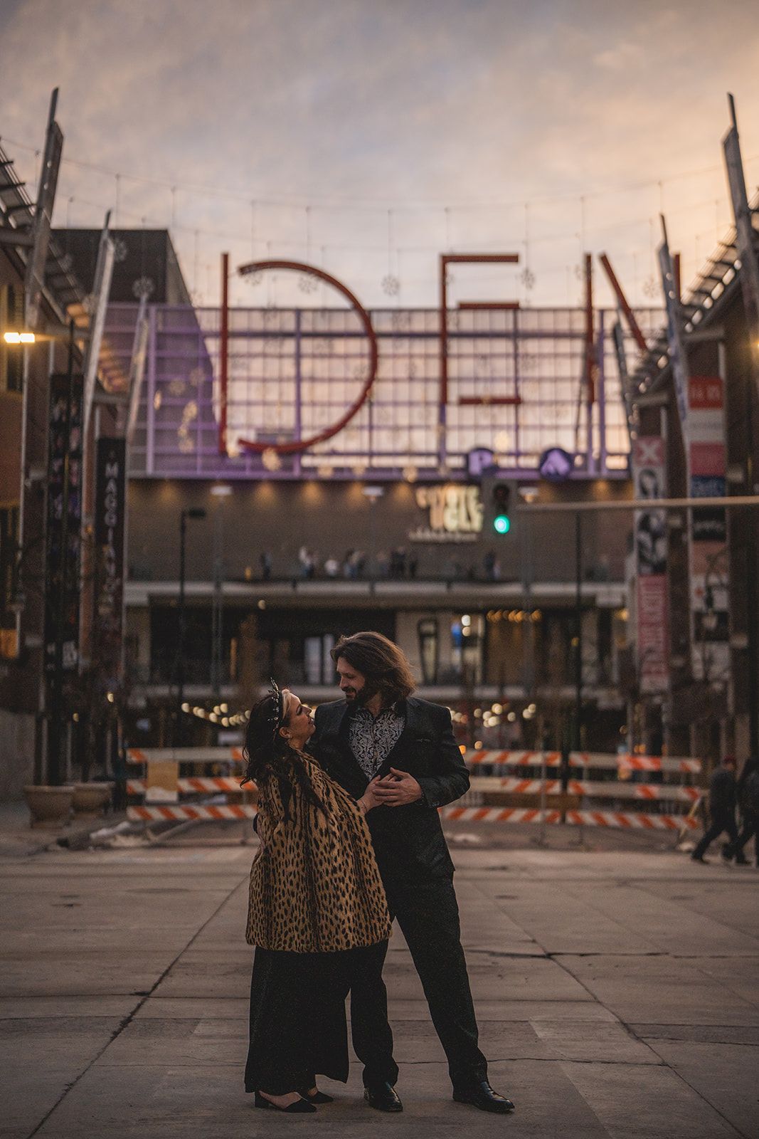 Couple embracing at dusk in front of illuminated DEN sign, woman in leopard coat, urban setting.