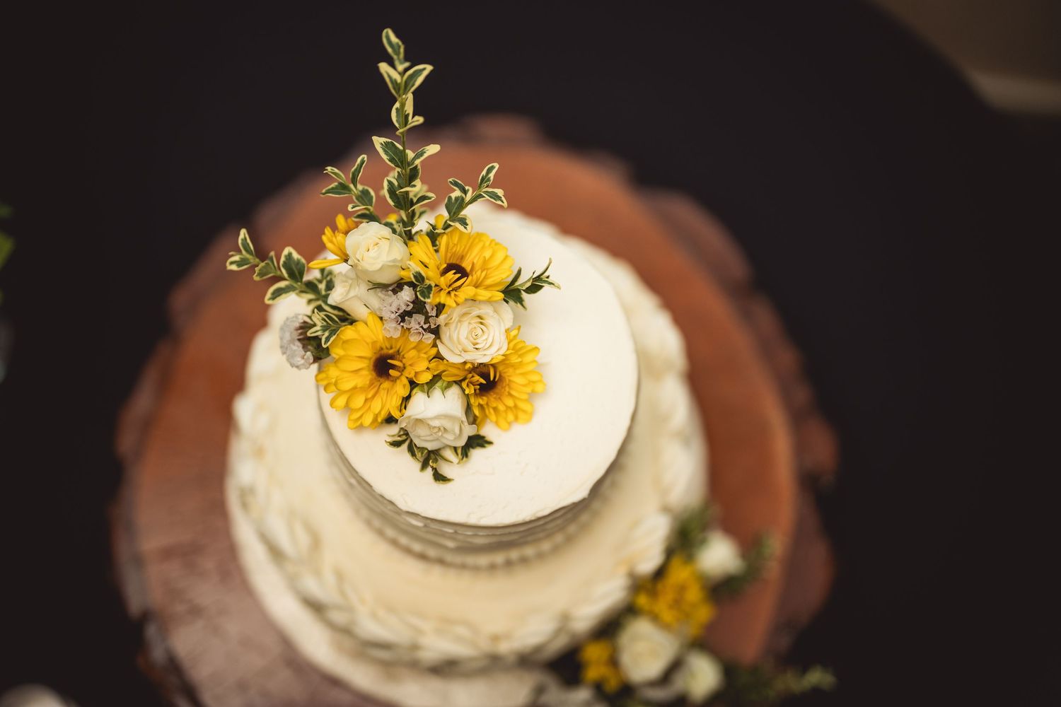 White wedding cake decorated with fresh yellow and white flowers on a dark wooden table.
