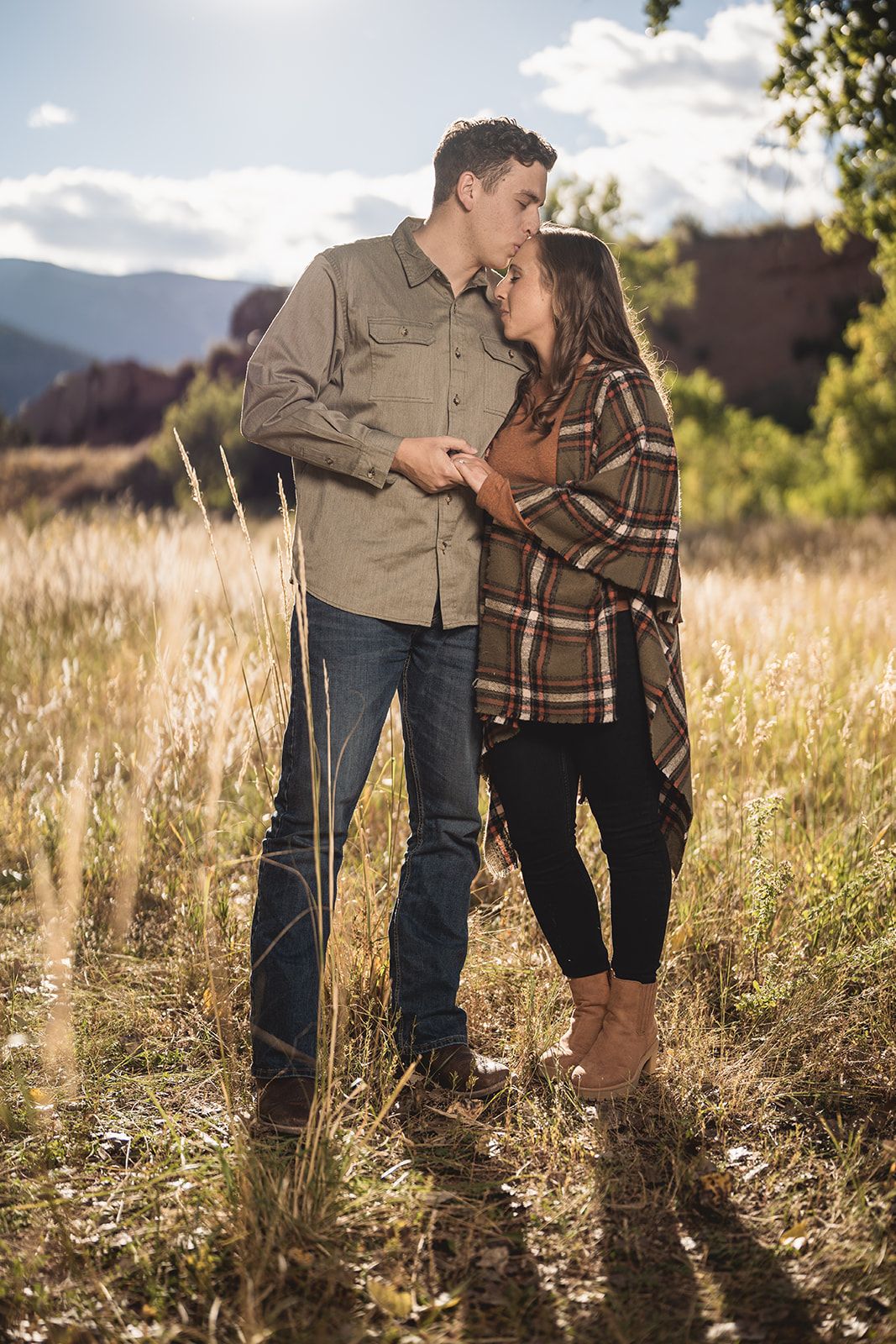 Couple in casual clothing shares intimate moment standing in tall grass with mountains in background.
