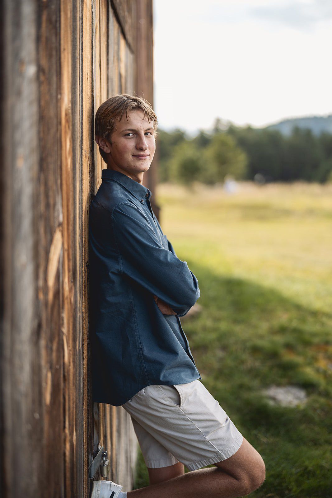 Young man in blue shirt and khaki shorts leaning against rustic wooden barn wall with green field backdrop.