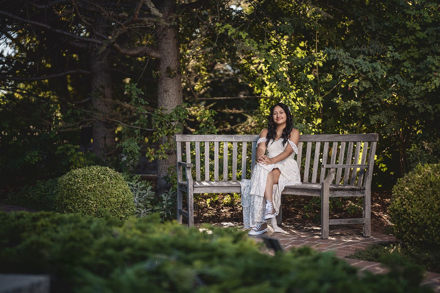 Woman in white dress sitting on wooden garden bench surrounded by lush green trees and shrubs in sunlight.
