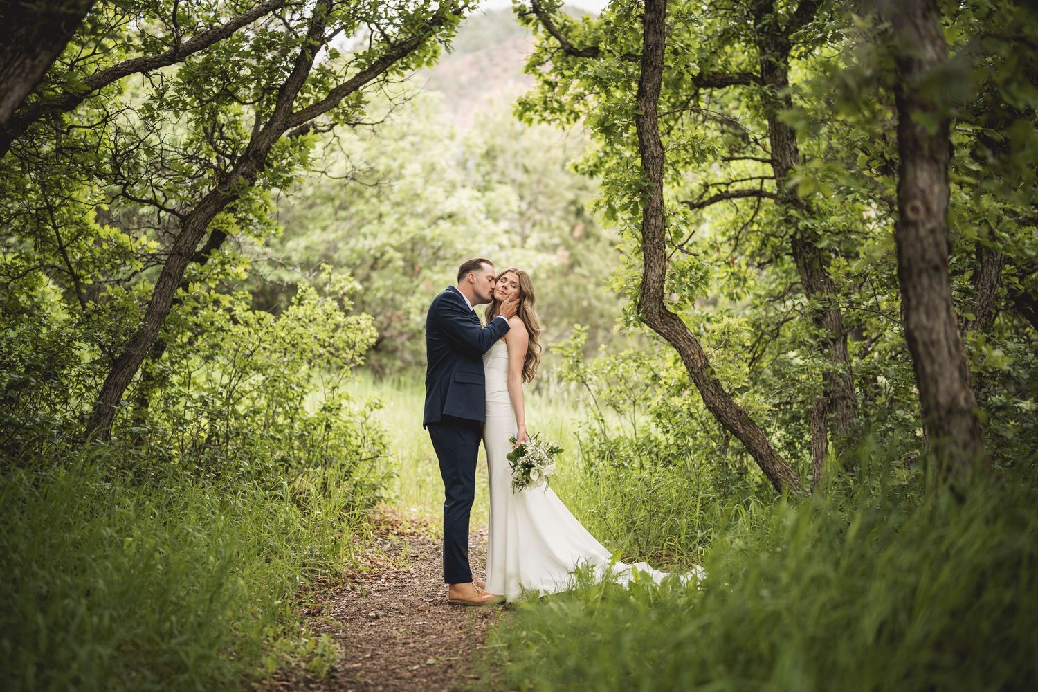 Couple shares intimate moment along wooded trail during outdoor wedding photoshoot.