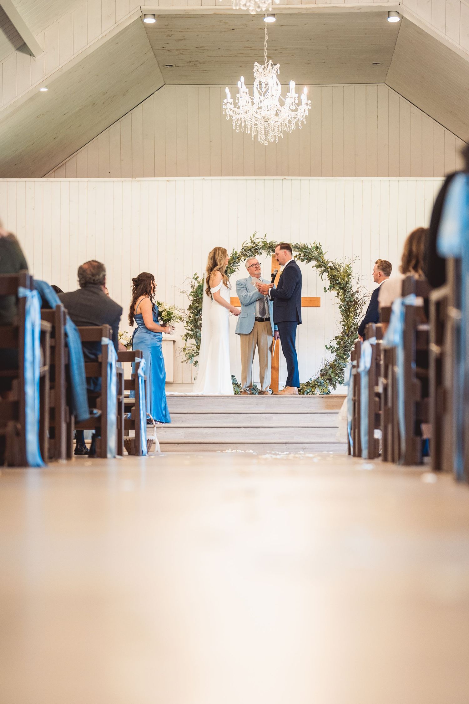 Wedding ceremony under crystal chandelier with floral arch and wooden church pews.