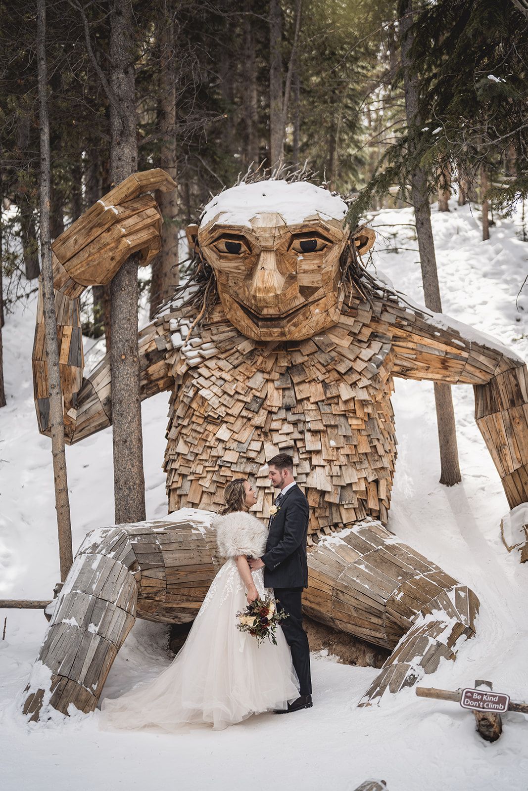 Bride and groom embrace before a giant wooden troll sculpture in a snowy forest winter wedding setting.