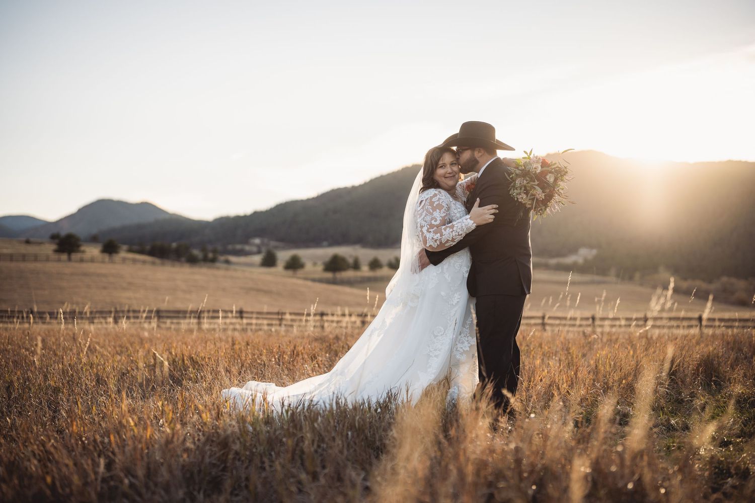 Bride and groom embrace in golden field at sunset with mountain backdrop, bride in lace gown, groom in black cowboy hat.