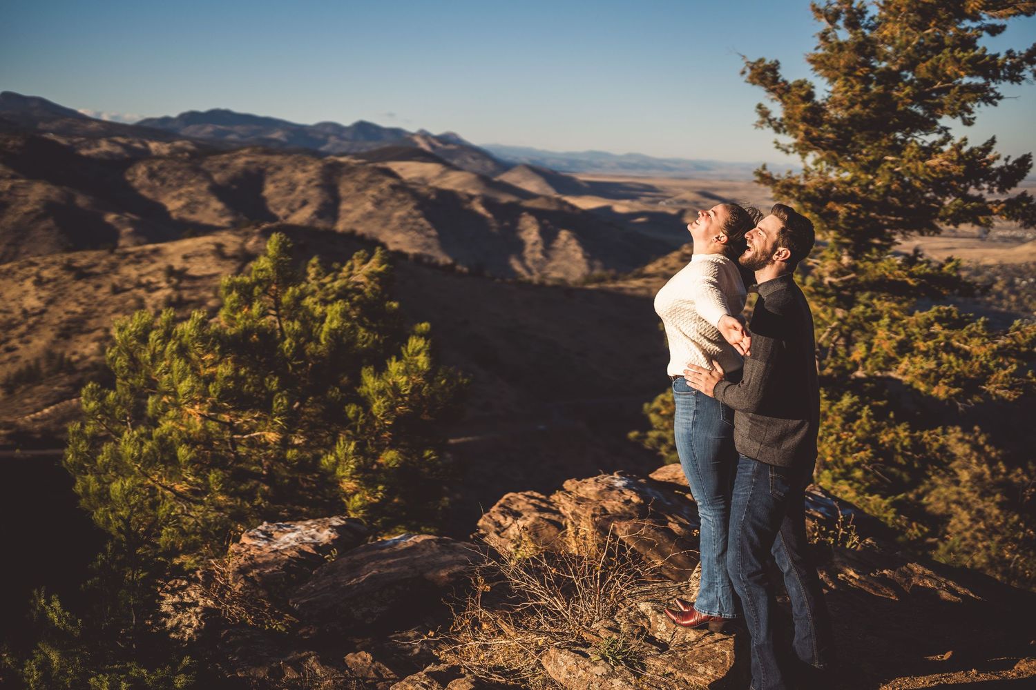 Two men embrace on a rocky mountain overlook surrounded by pine trees with sweeping desert valley views.