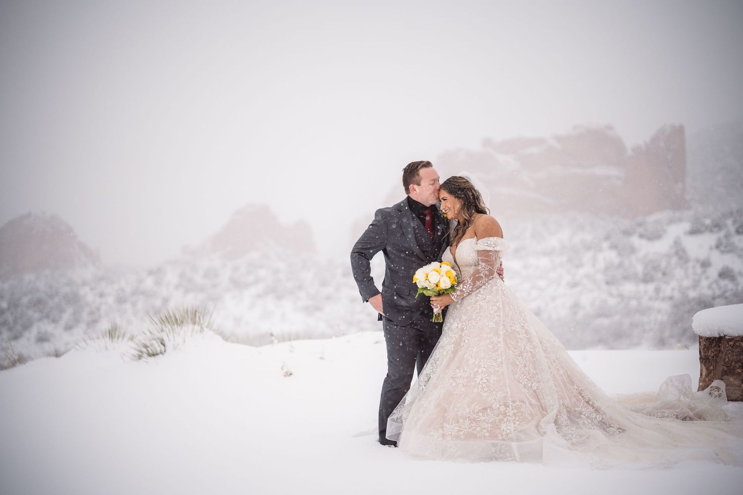 Newlyweds share intimate moment against snowy mountain backdrop in formal winter attire.