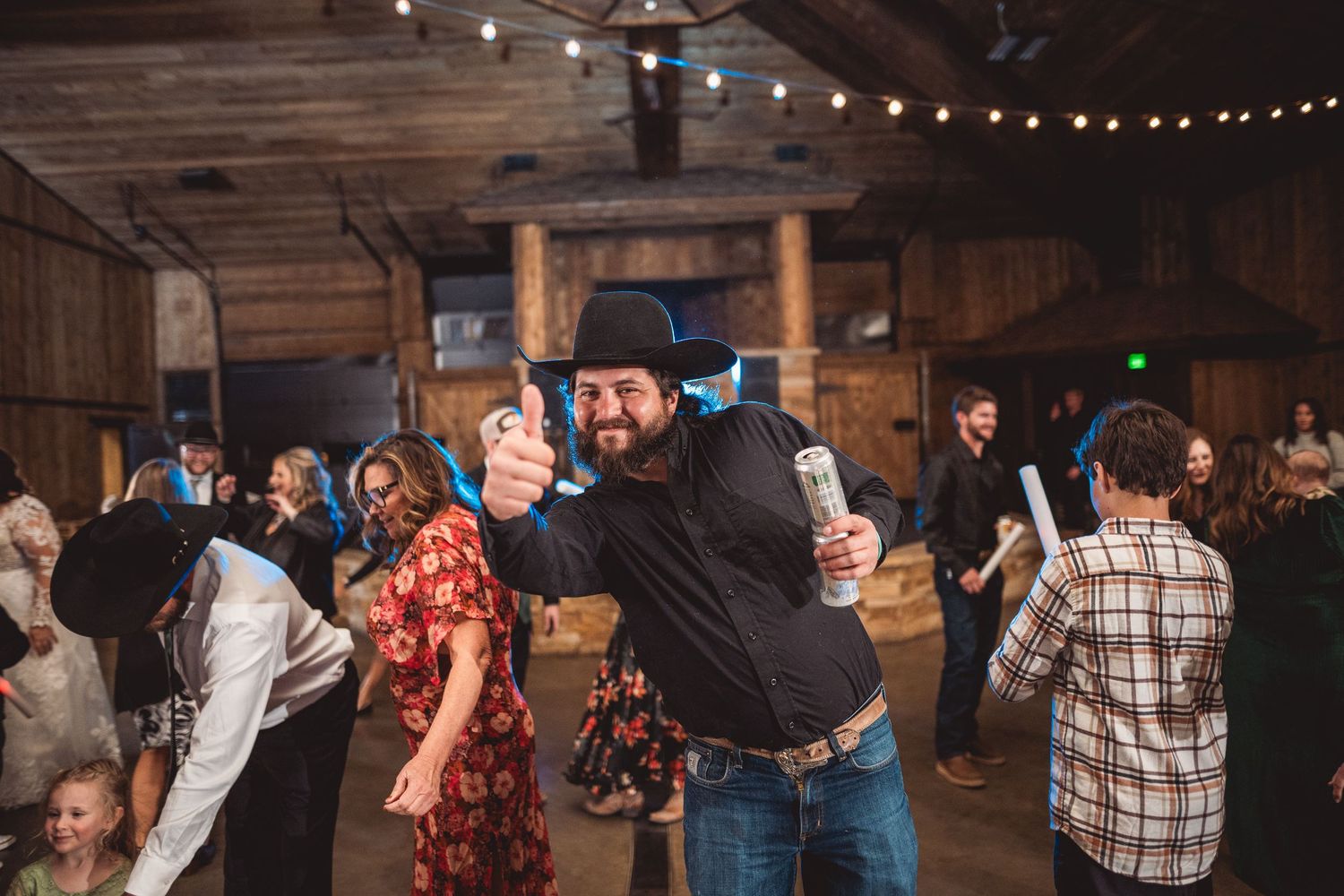 People dancing and celebrating in a rustic wooden barn with string lights overhead.