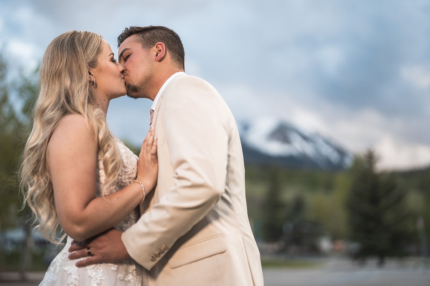 Bride and groom share a romantic kiss outdoors with snow-capped mountains and evergreen trees in the background.