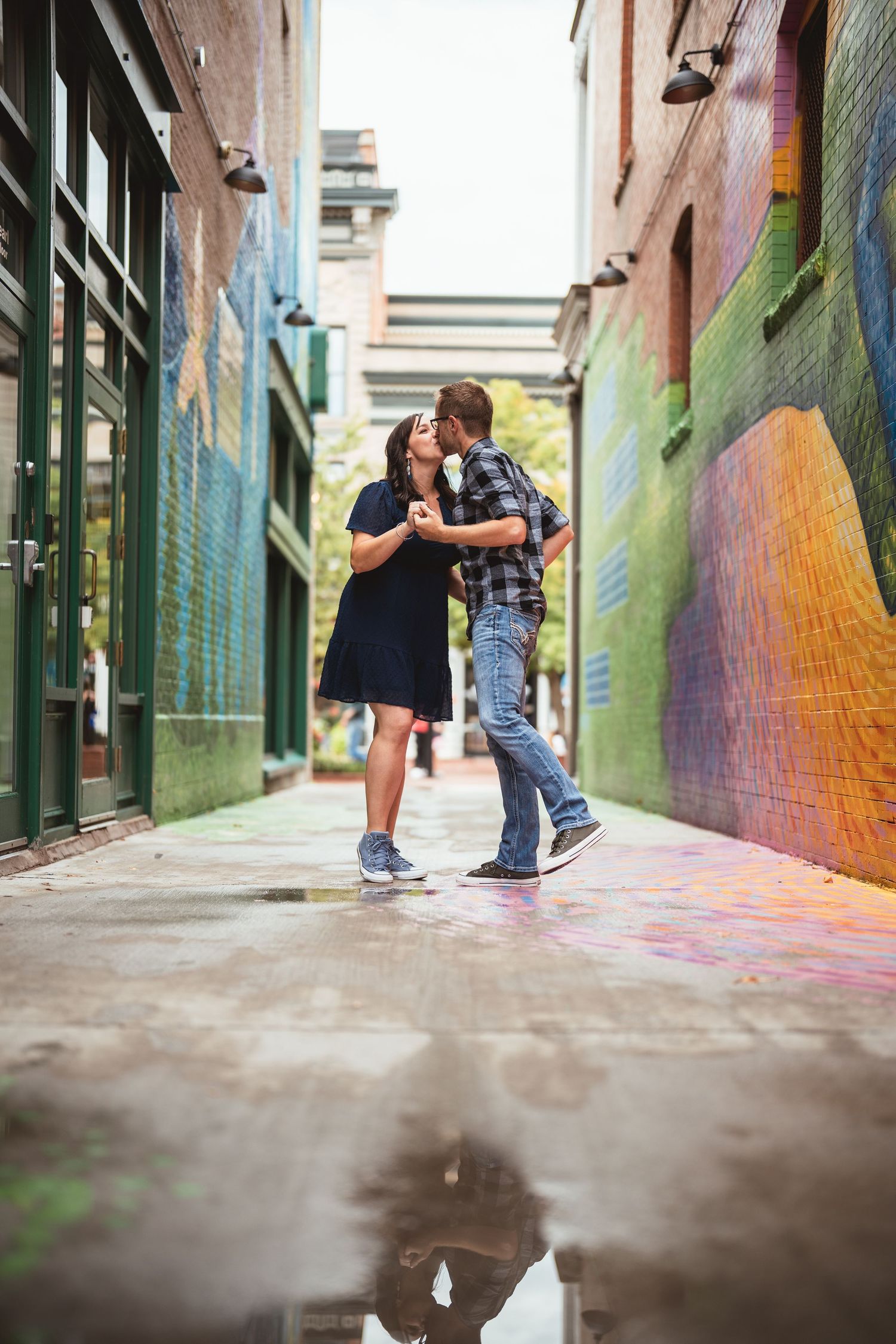 Couple sharing a romantic kiss in a colorful urban alley with vibrant murals and wet pavement reflections.