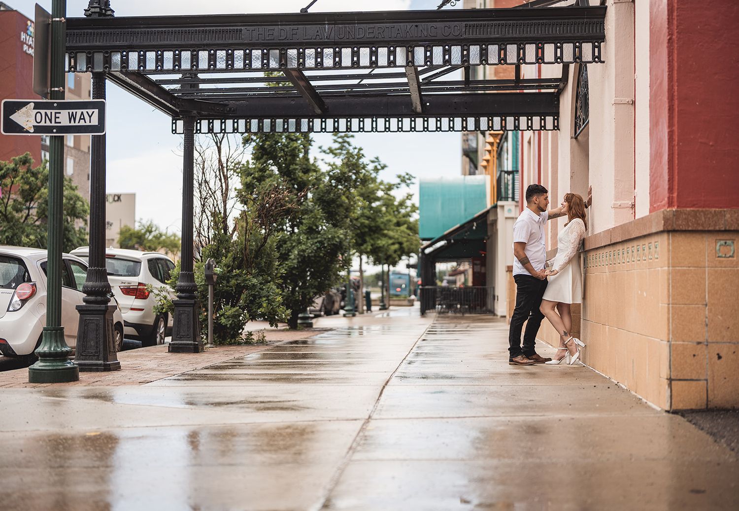 Couple sharing a romantic moment on a rainy urban sidewalk near a one-way sign and charming storefronts.