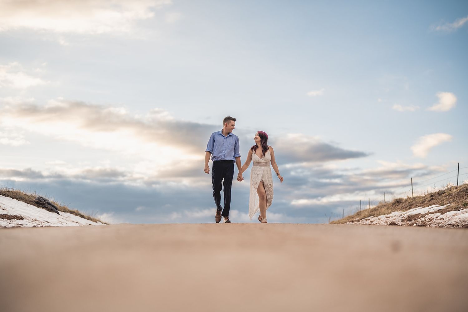 Couple holding hands walking on sandy beach dunes under blue sky at sunset engagement photo.