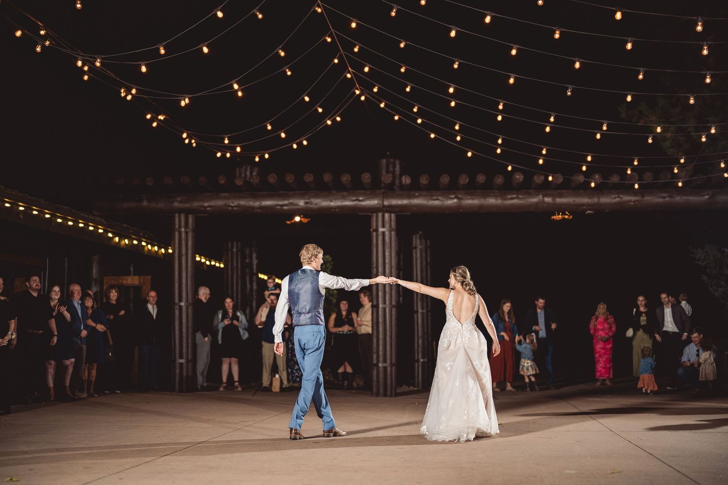 Bride and groom share first dance under string lights at outdoor wedding reception with guests watching.