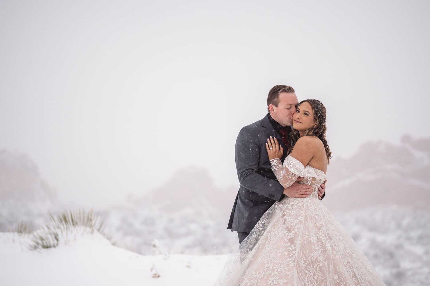 Couple shares romantic kiss in snowy winter wedding, bride in lace gown, groom in gray suit, red rocks backdrop.