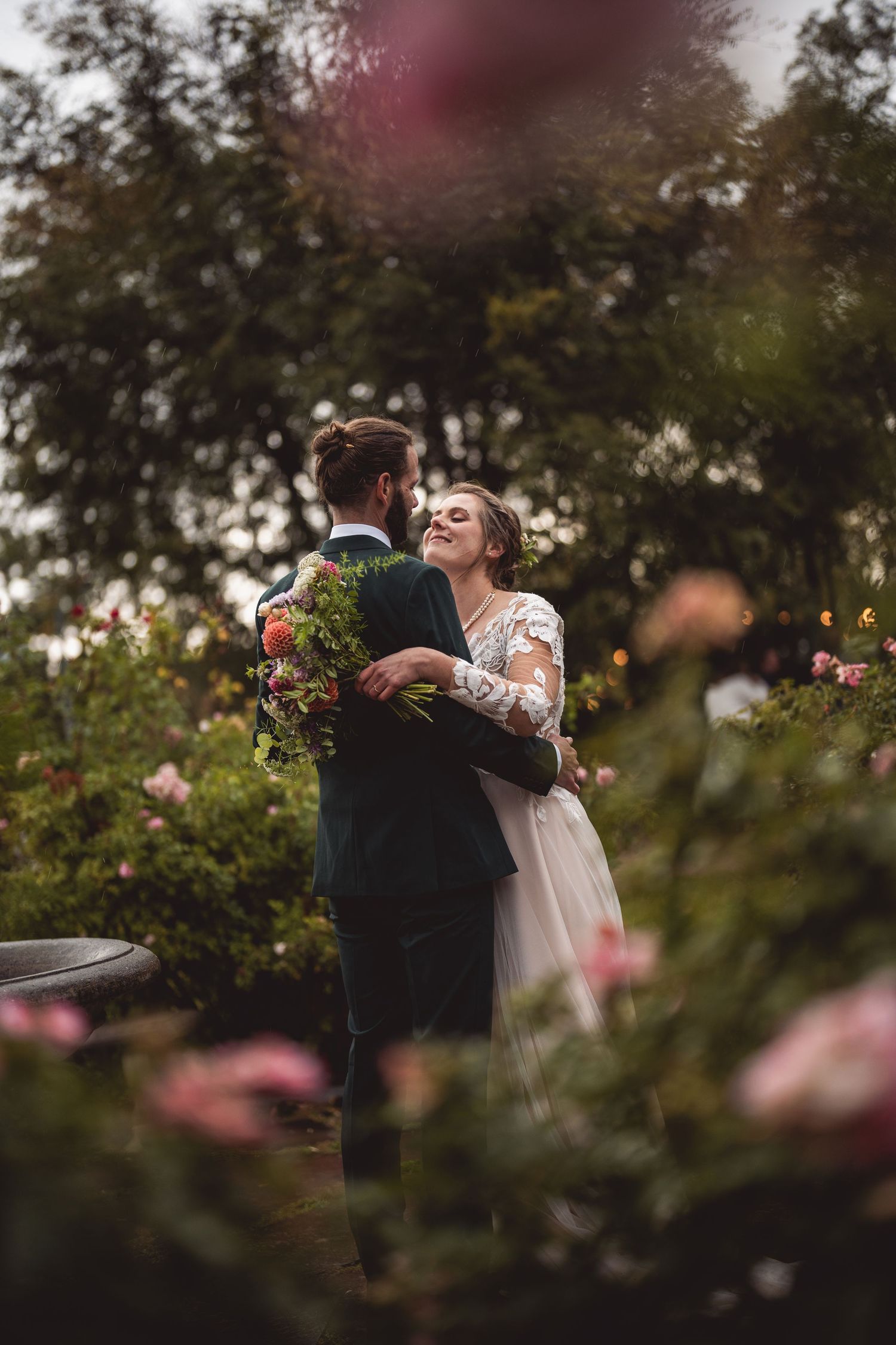 Bride and groom embrace in a lush rose garden, bride smiling joyfully in lace gown holding bouquet.