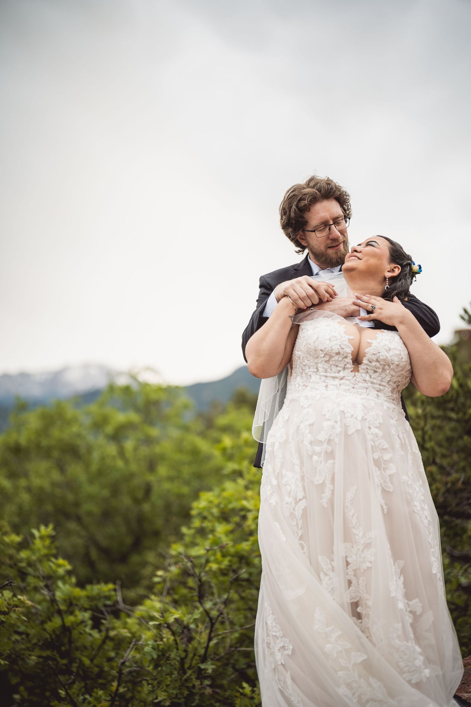 Groom embraces bride from behind in lace gown with mountain landscape backdrop on their wedding day.