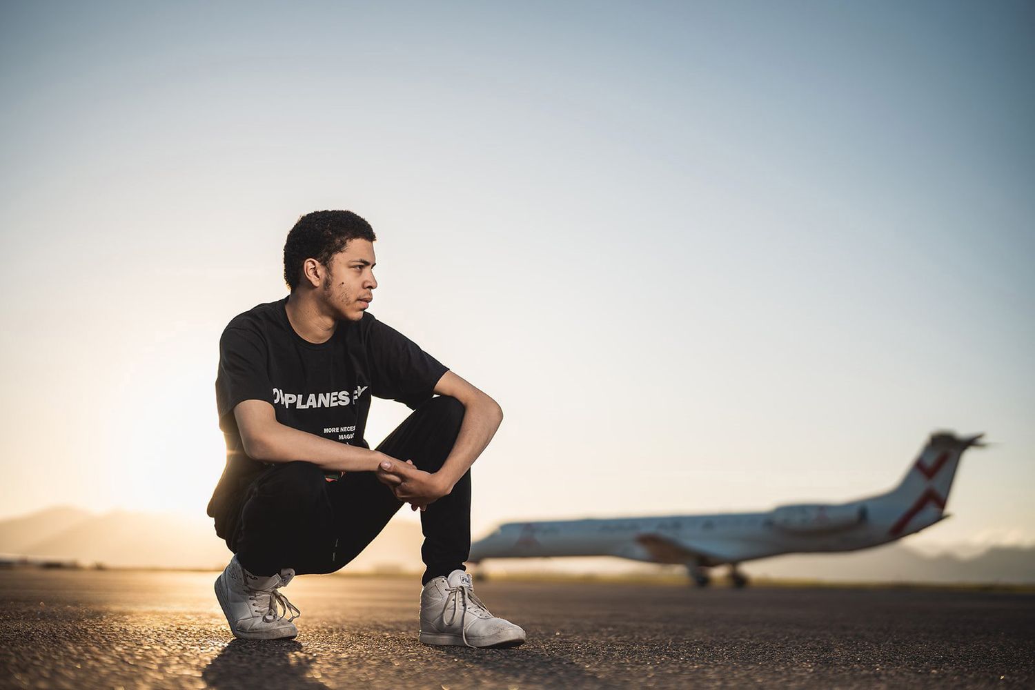 Young man in black t-shirt crouching on airport tarmac with private jet at sunset.