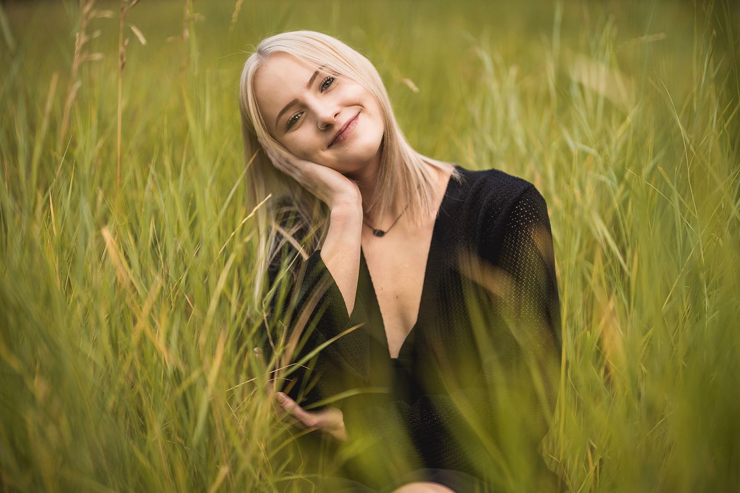 Smiling blonde woman in black top relaxing in tall green grass field outdoors.