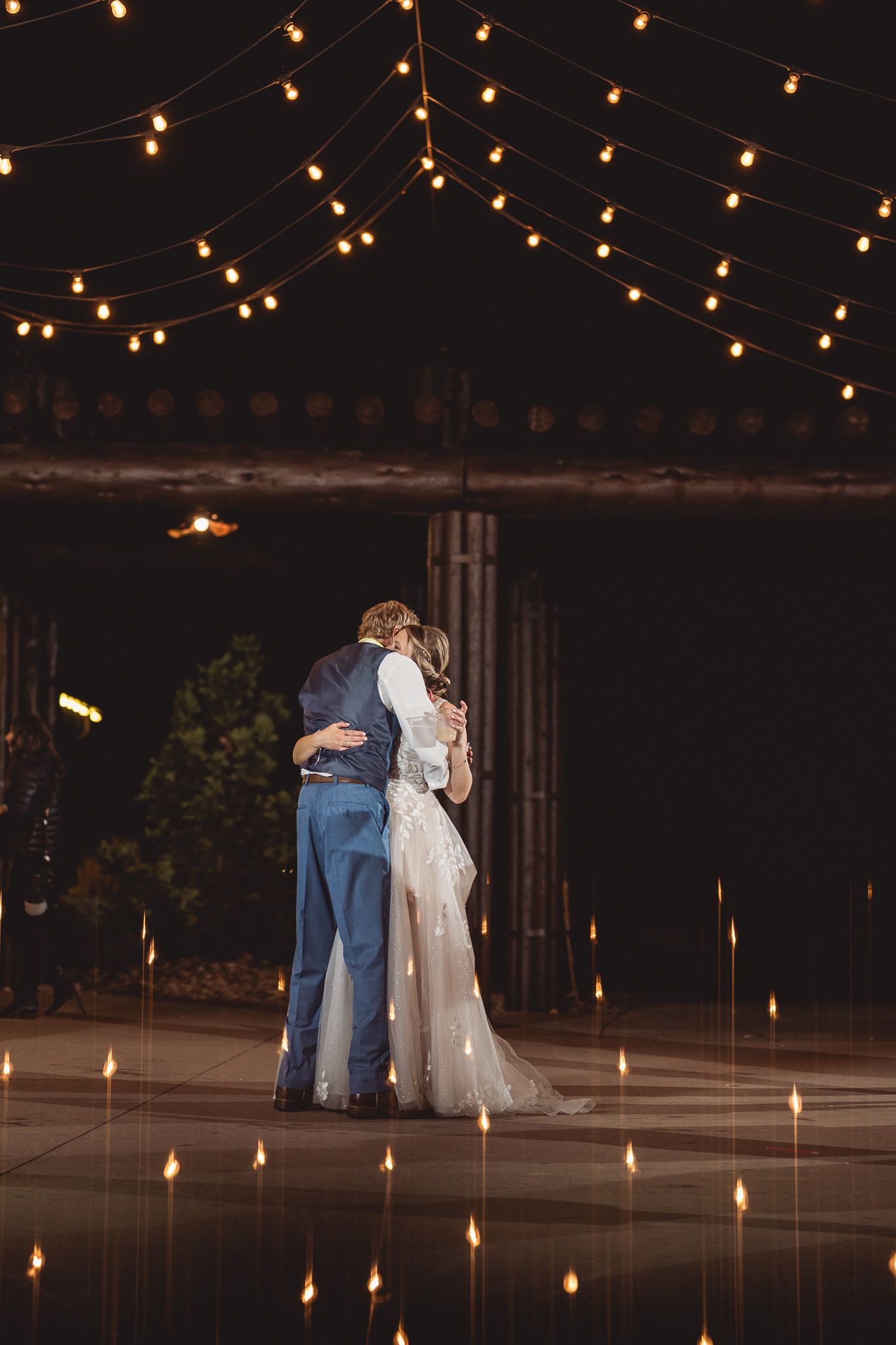 Bride and groom share first dance under warm string lights at night on a reflective outdoor dance floor.