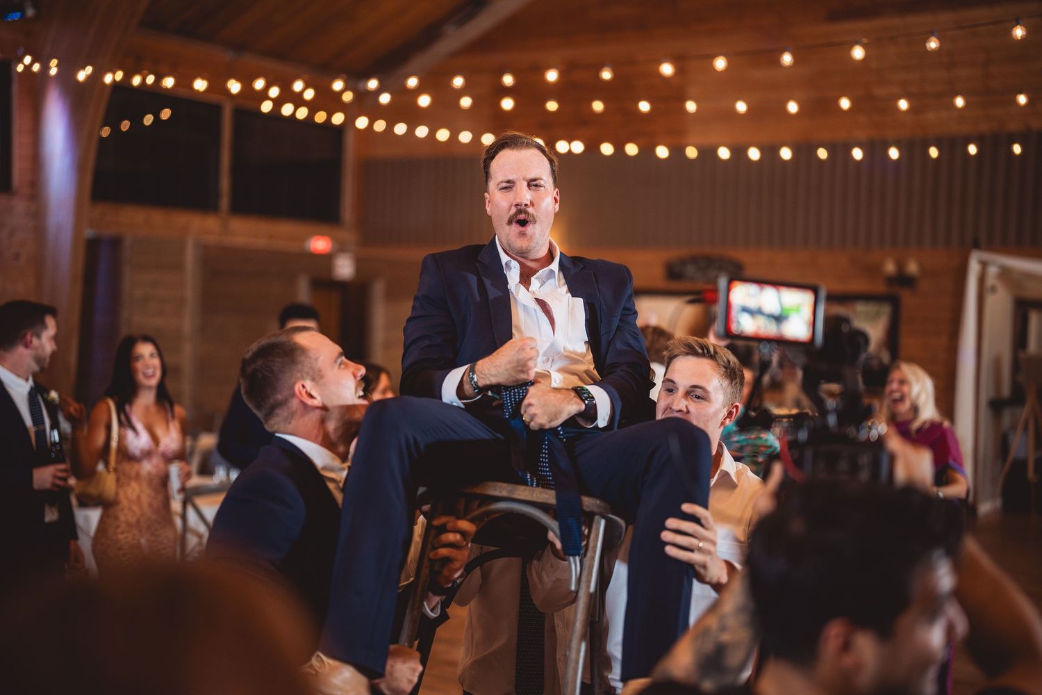 String lights create a warm ambiance in an indoor reception venue with exposed wooden beams.