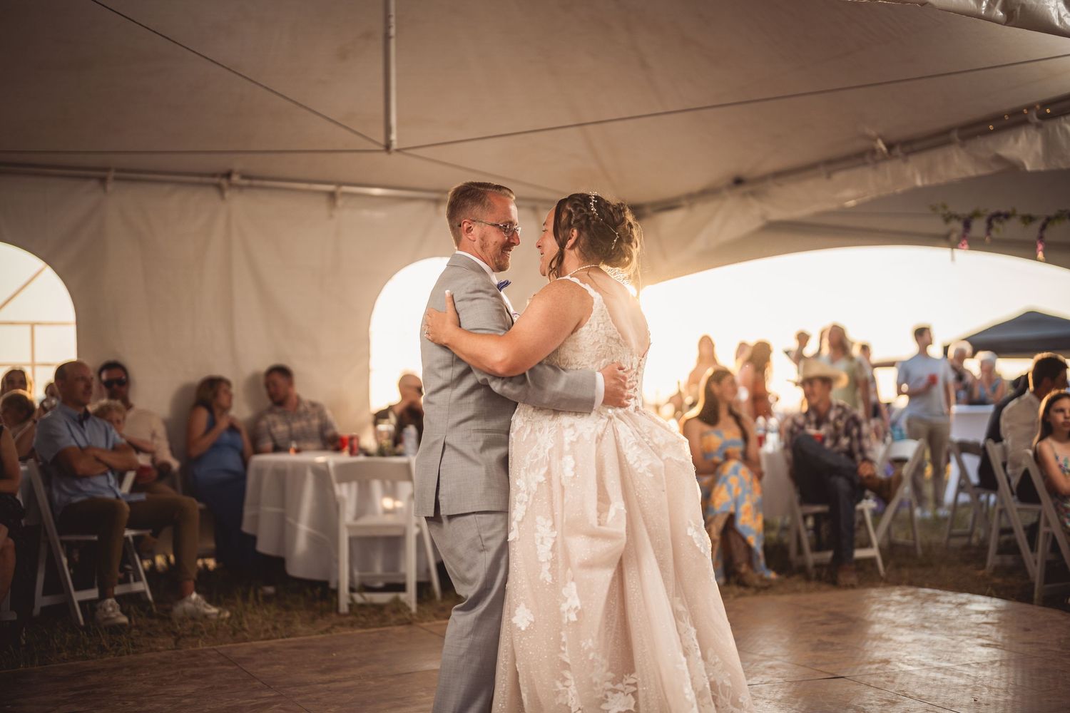 Couple dances together during wedding reception under white tent with guests seated at tables.
