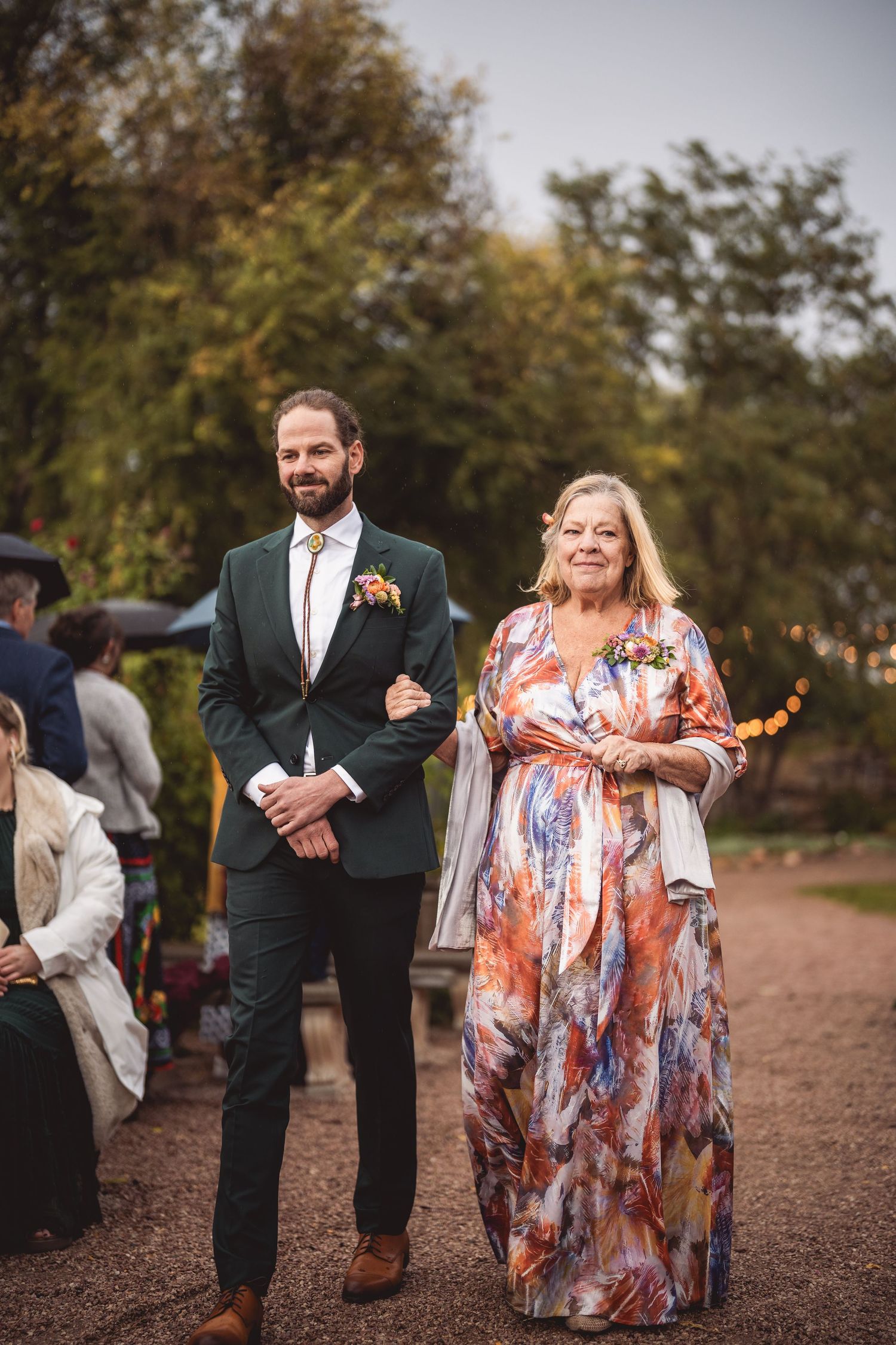 Wedding party members walk down a gravel path lined with trees and string lights.