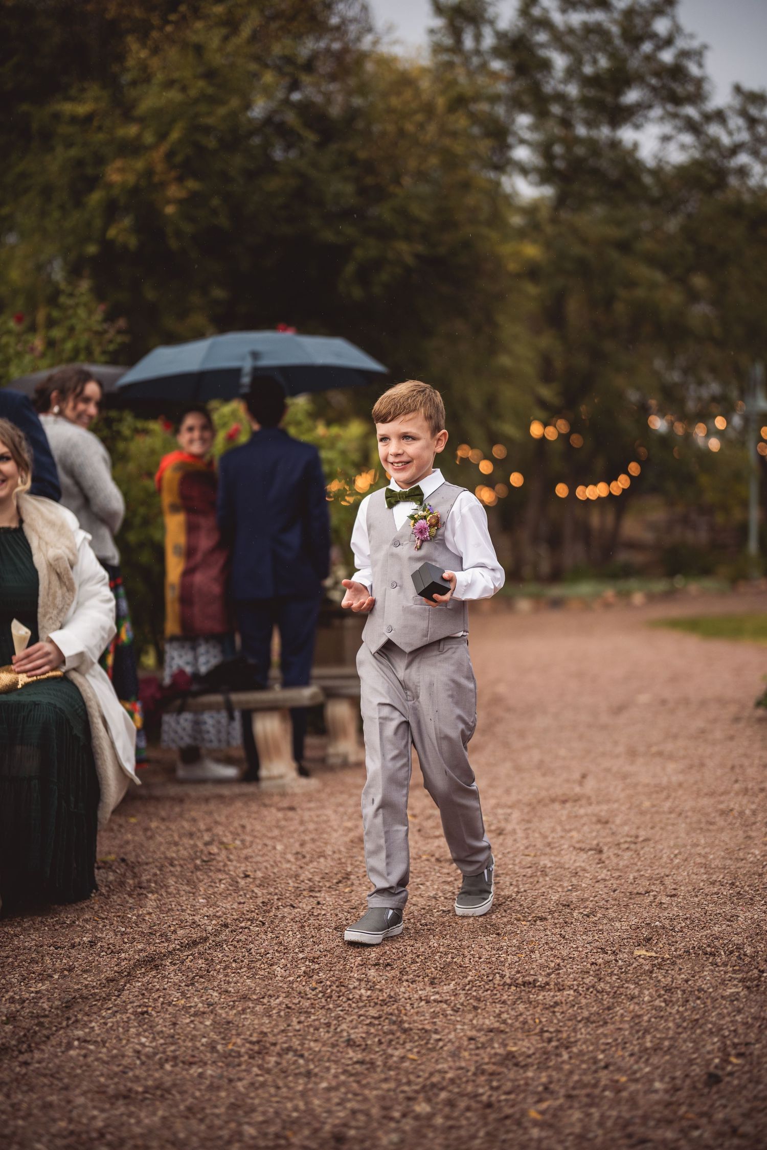 Children in formal attire playfully run down a gravel path at outdoor wedding.