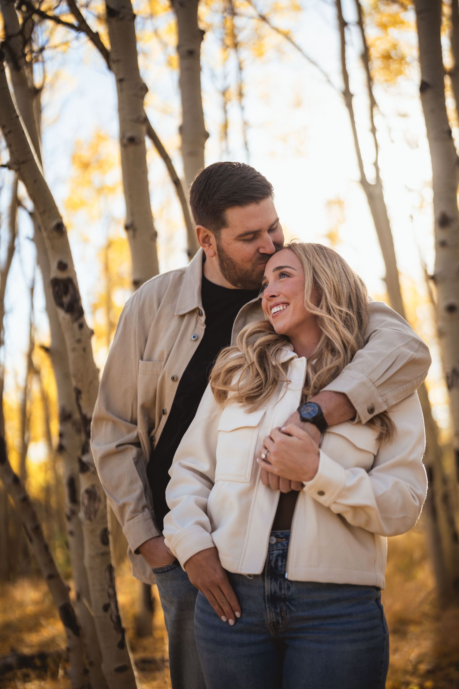 Couple sharing a joyful moment among autumn birch trees in matching beige jackets.