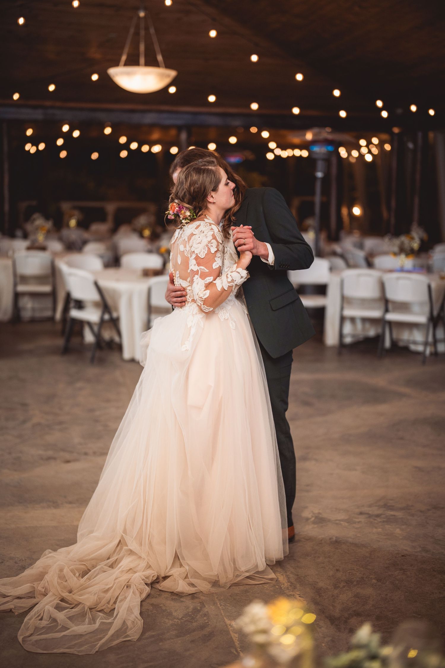Romantic dance moment in a rustic reception hall with flowing white dress and string lights.