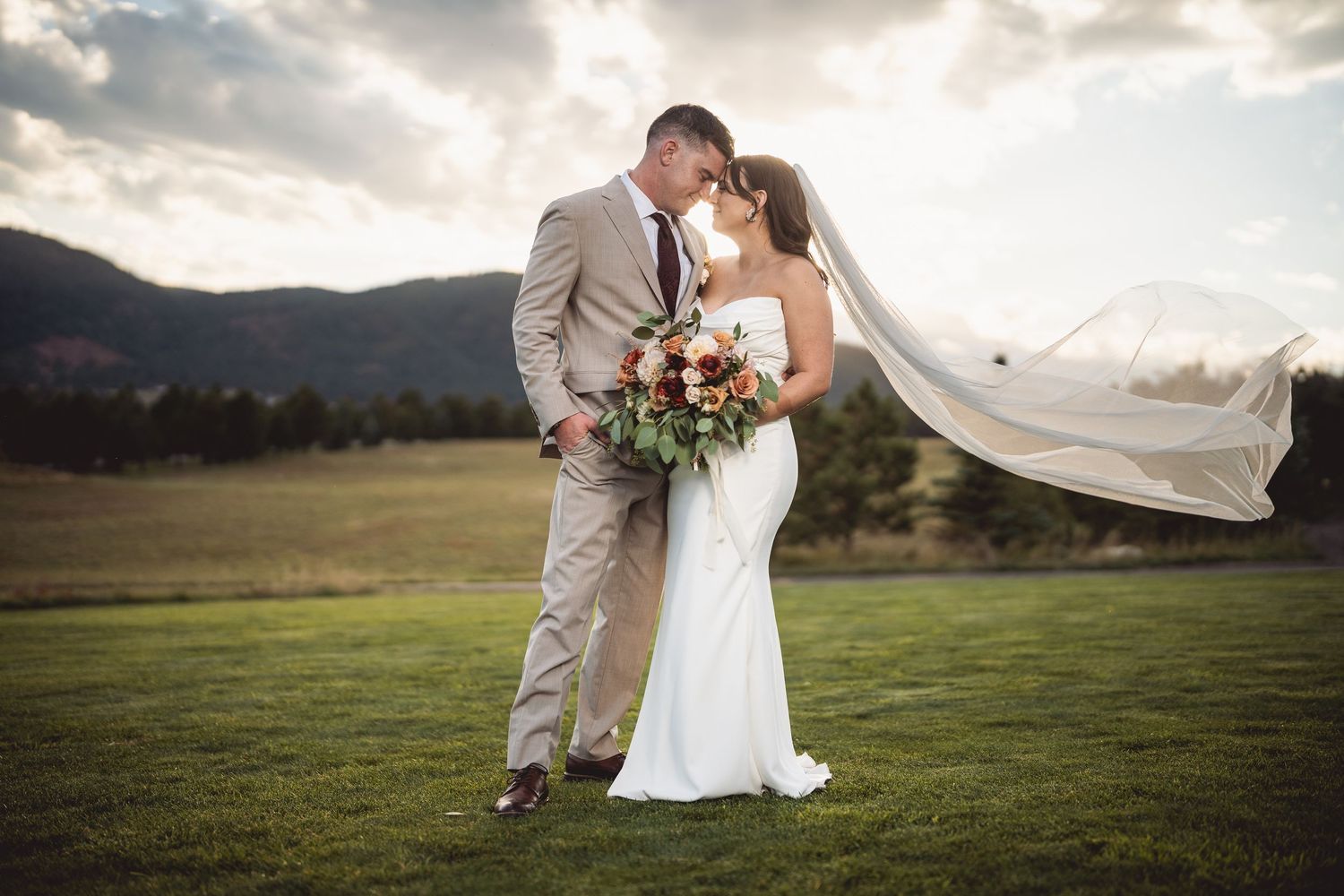 Bride and groom kissing on green lawn with veil blowing in wind and mountain backdrop at sunset.