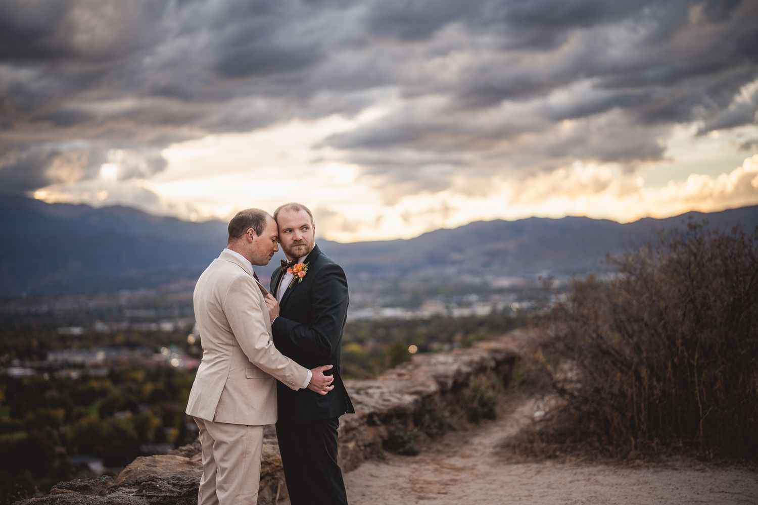 Two grooms share an intimate moment on a hilltop at sunset with dramatic cloudy skies and mountains.
