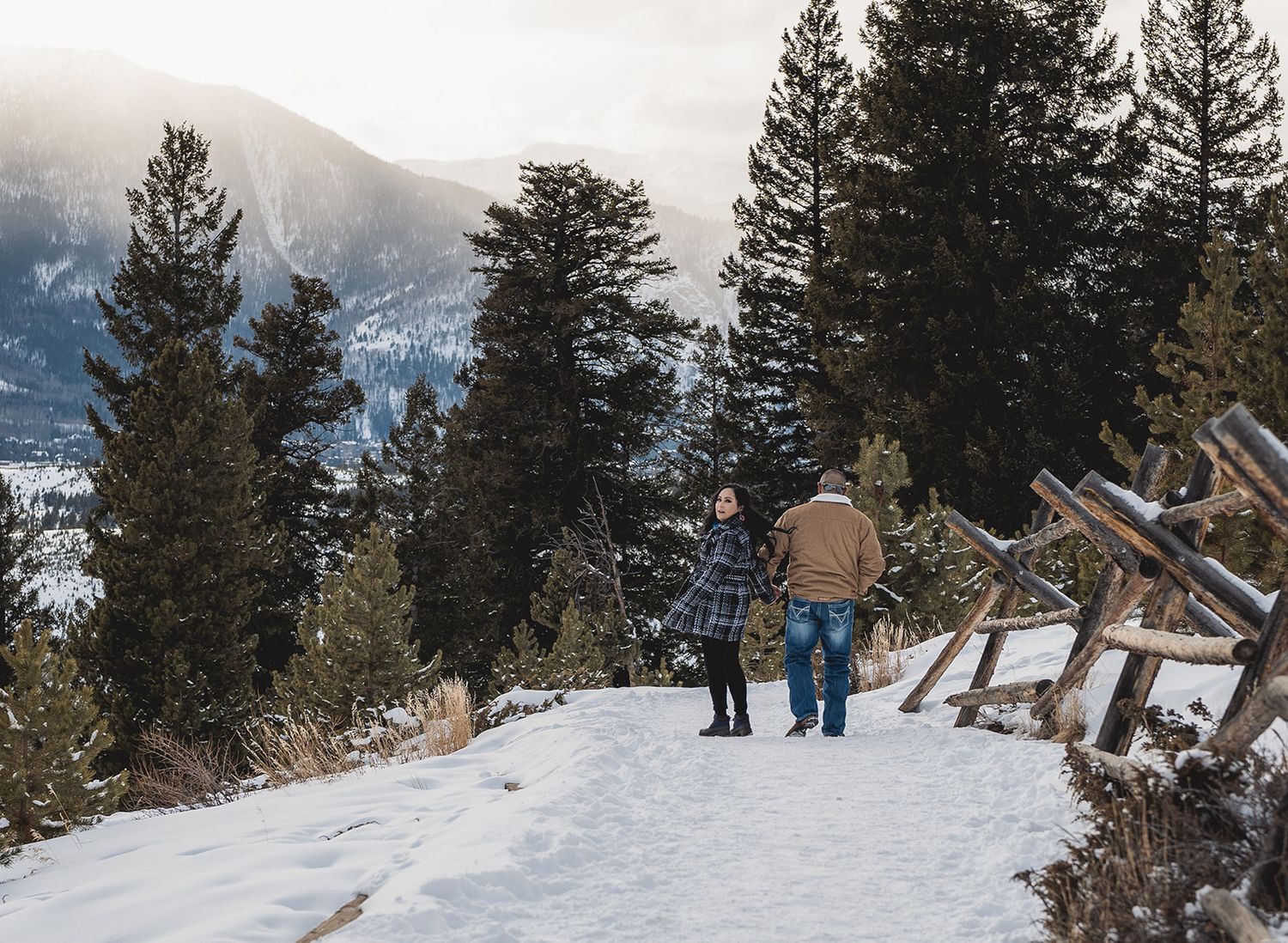 Couple walking on a snowy mountain trail surrounded by pine trees and scenic winter landscape.