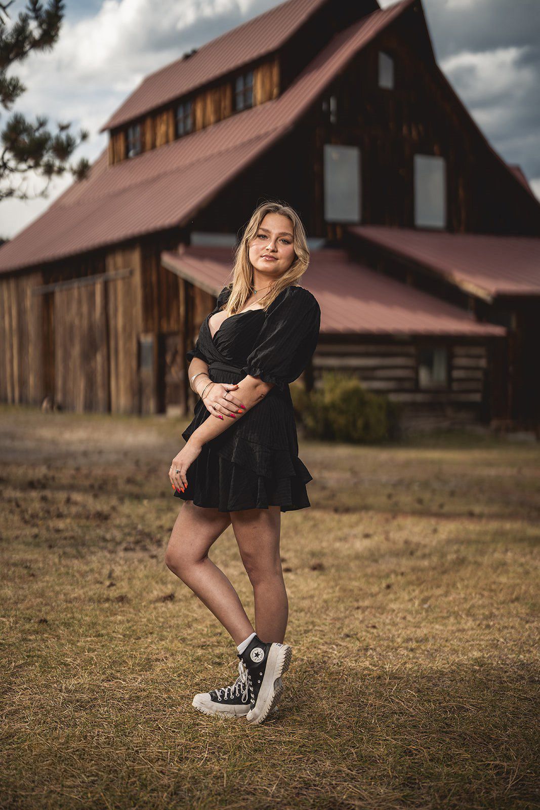 Blonde woman in black mini dress and Converse sneakers posing in front of a rustic wooden barn.