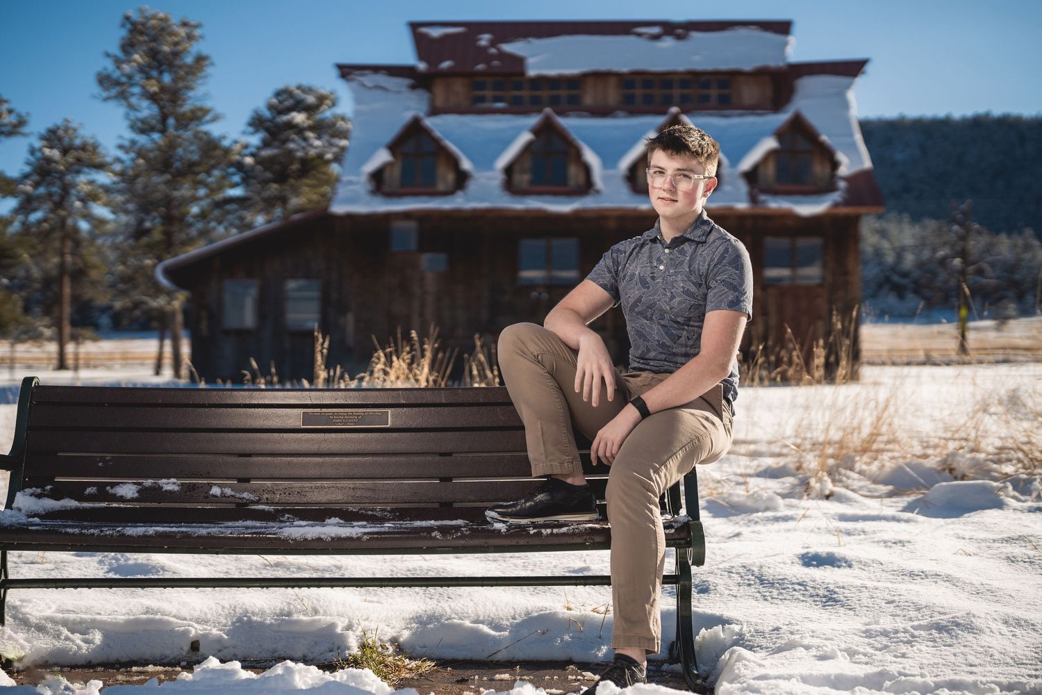 Teen boy sitting on a bench in a snowy winter landscape with a log cabin in the background.