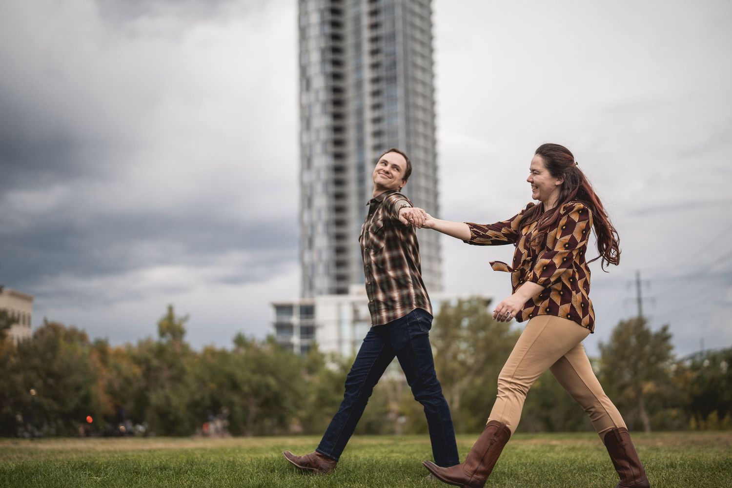 Couple holding hands walking in a park with a modern skyscraper and cloudy sky in the background.