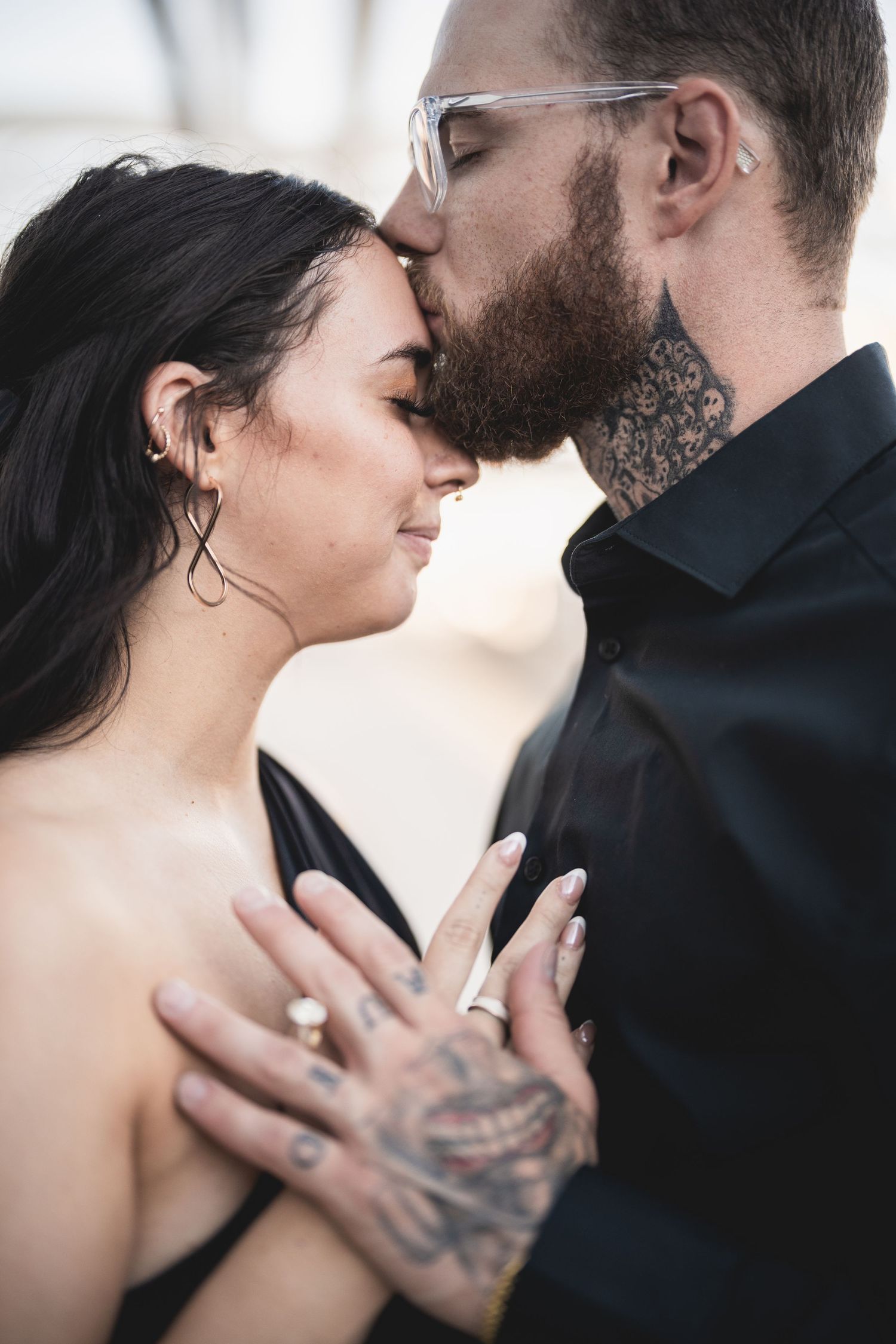 Tattooed couple sharing a romantic forehead kiss, woman wearing ring, man in black shirt with glasses and neck tattoos.