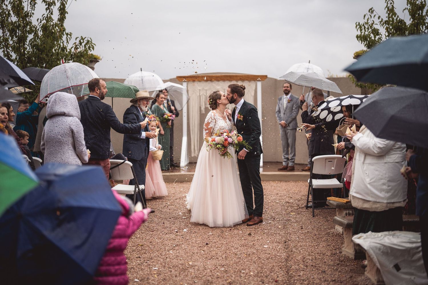 Wide shot of wedding celebration with attendees holding umbrellas in courtyard.