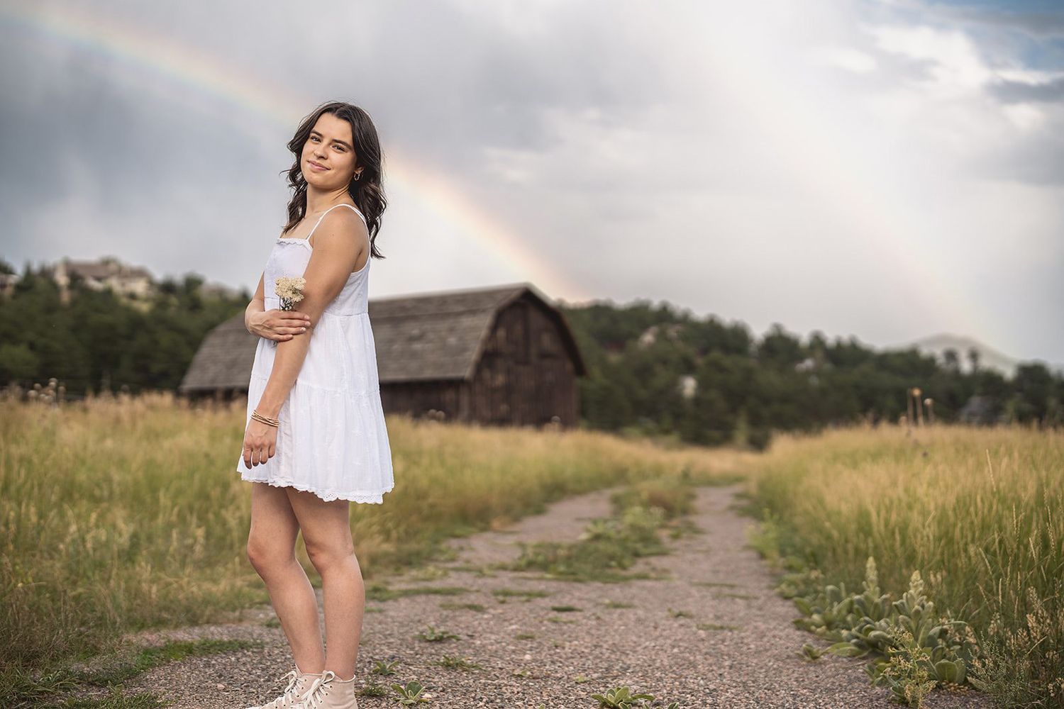 Woman in white dress standing on dirt road with rainbow and barn in background.