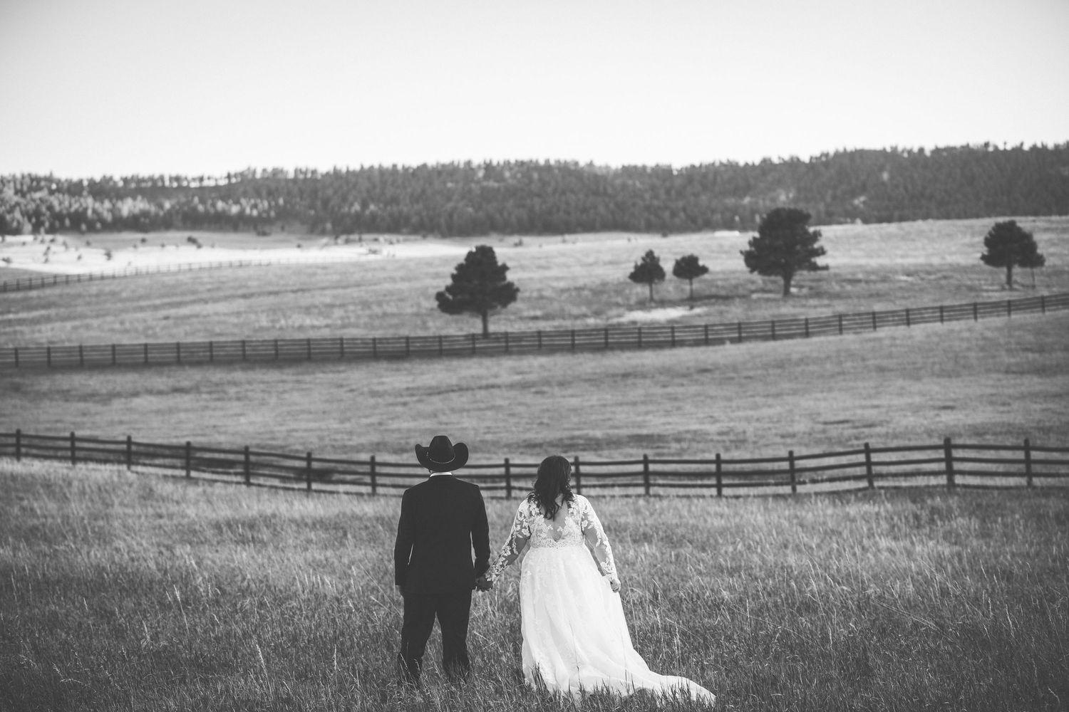 A couple stands in a rustic field with wooden fences and scattered trees against a misty mountain backdrop.