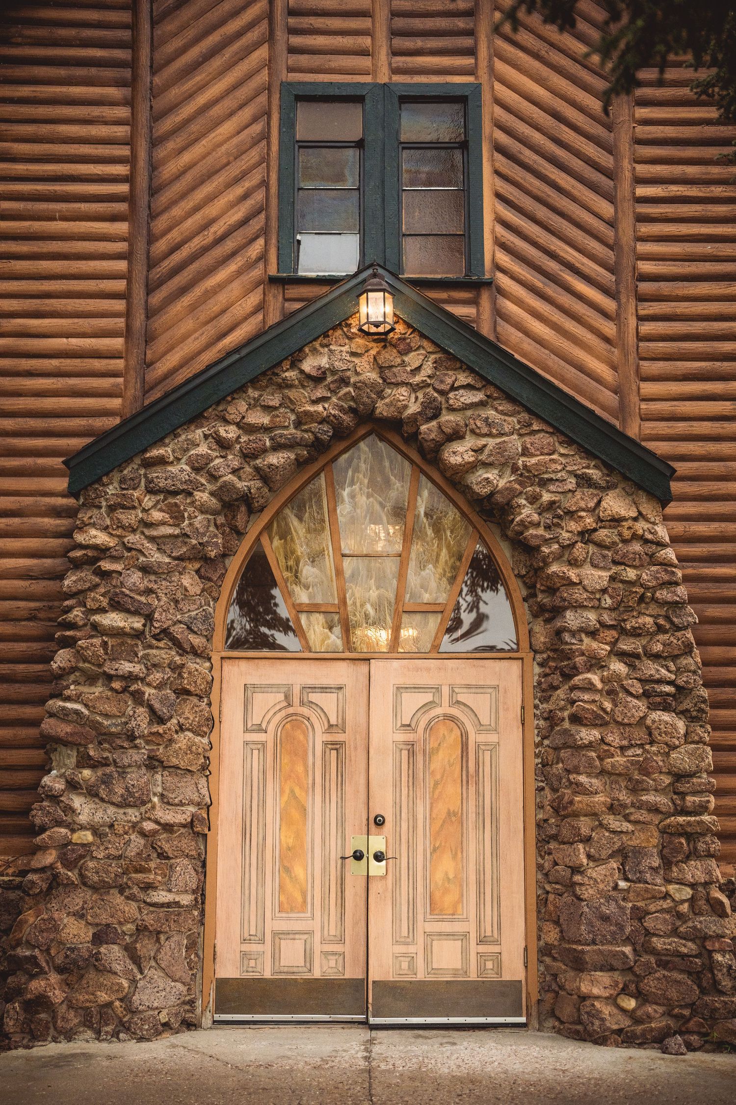 Rustic stone church entrance with Gothic arched doorway and wooden log siding illuminated by lantern light.