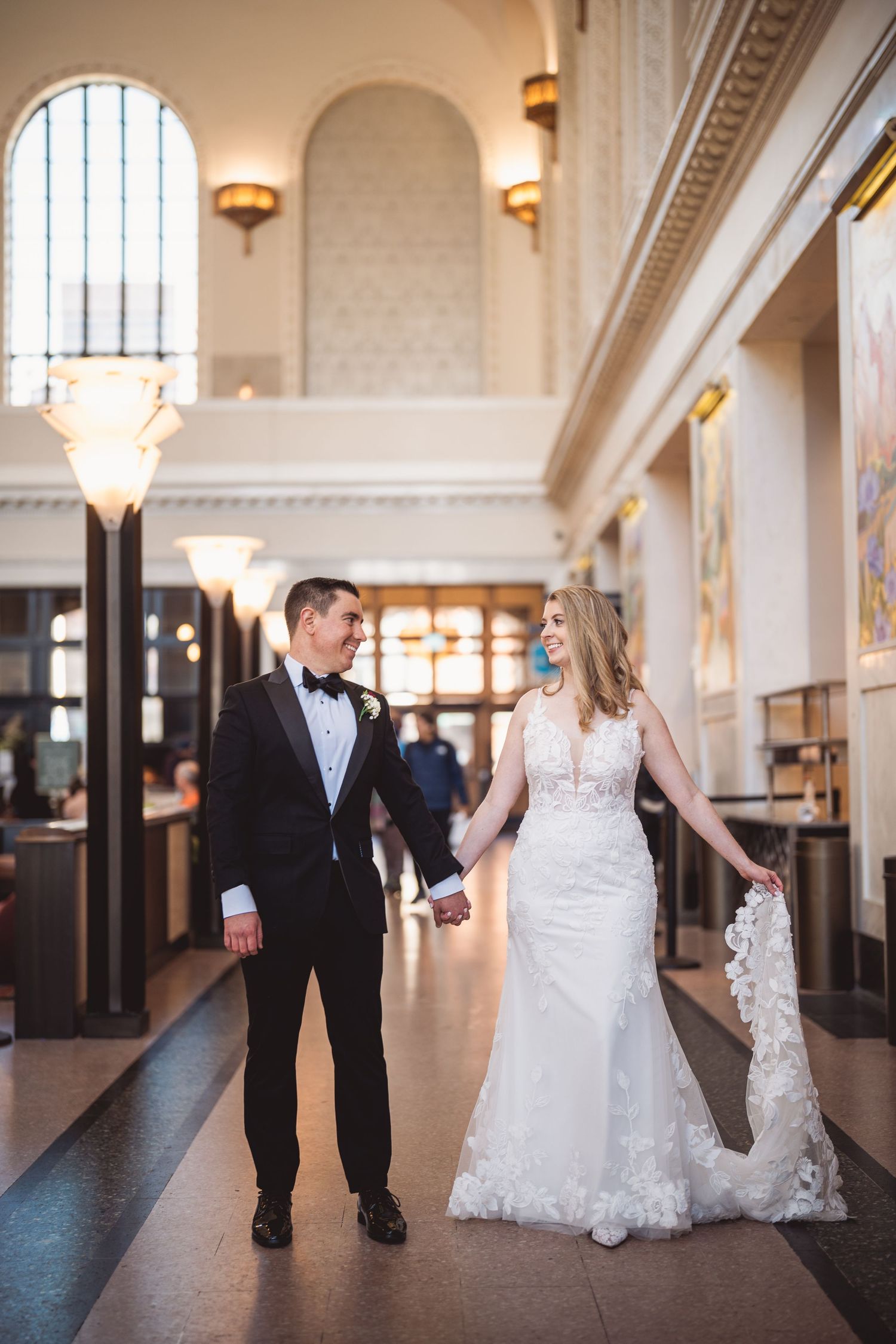 Newlyweds walking together in an elegant hallway with arched ceilings and chandeliers.