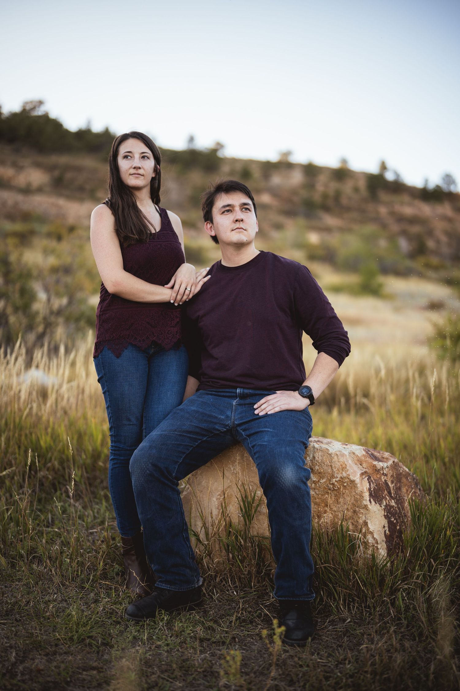 A couple sits together on a rock surrounded by tall grass in a natural outdoor setting.