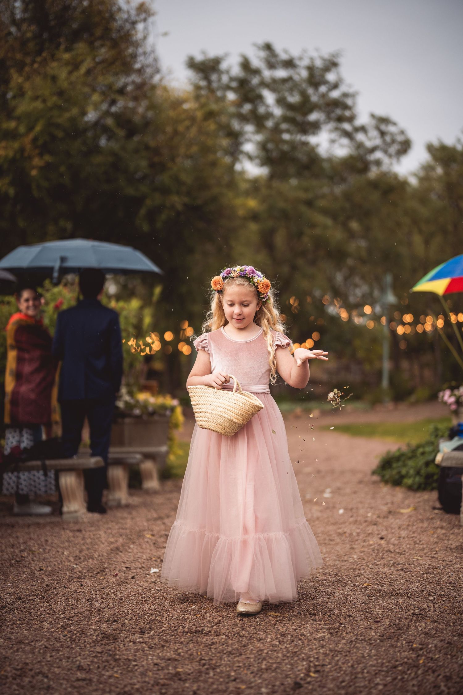 Young flower girl in pink tulle dress plays with flower petals on gravel path.