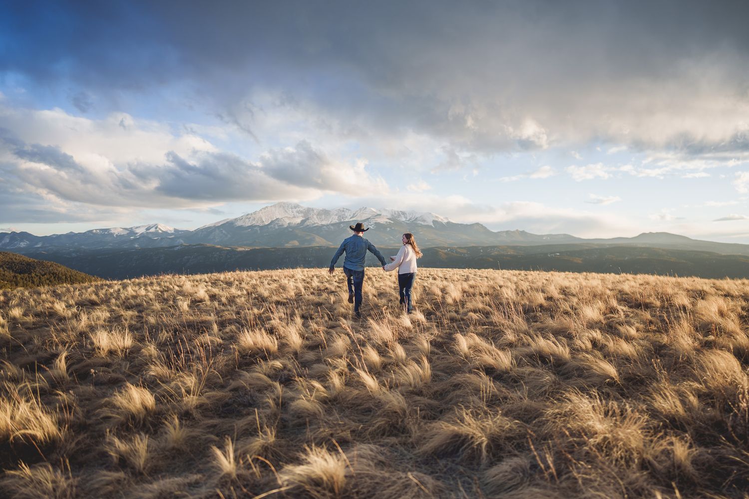 Wedding couple walking through golden grass field with mountain landscape in the background.