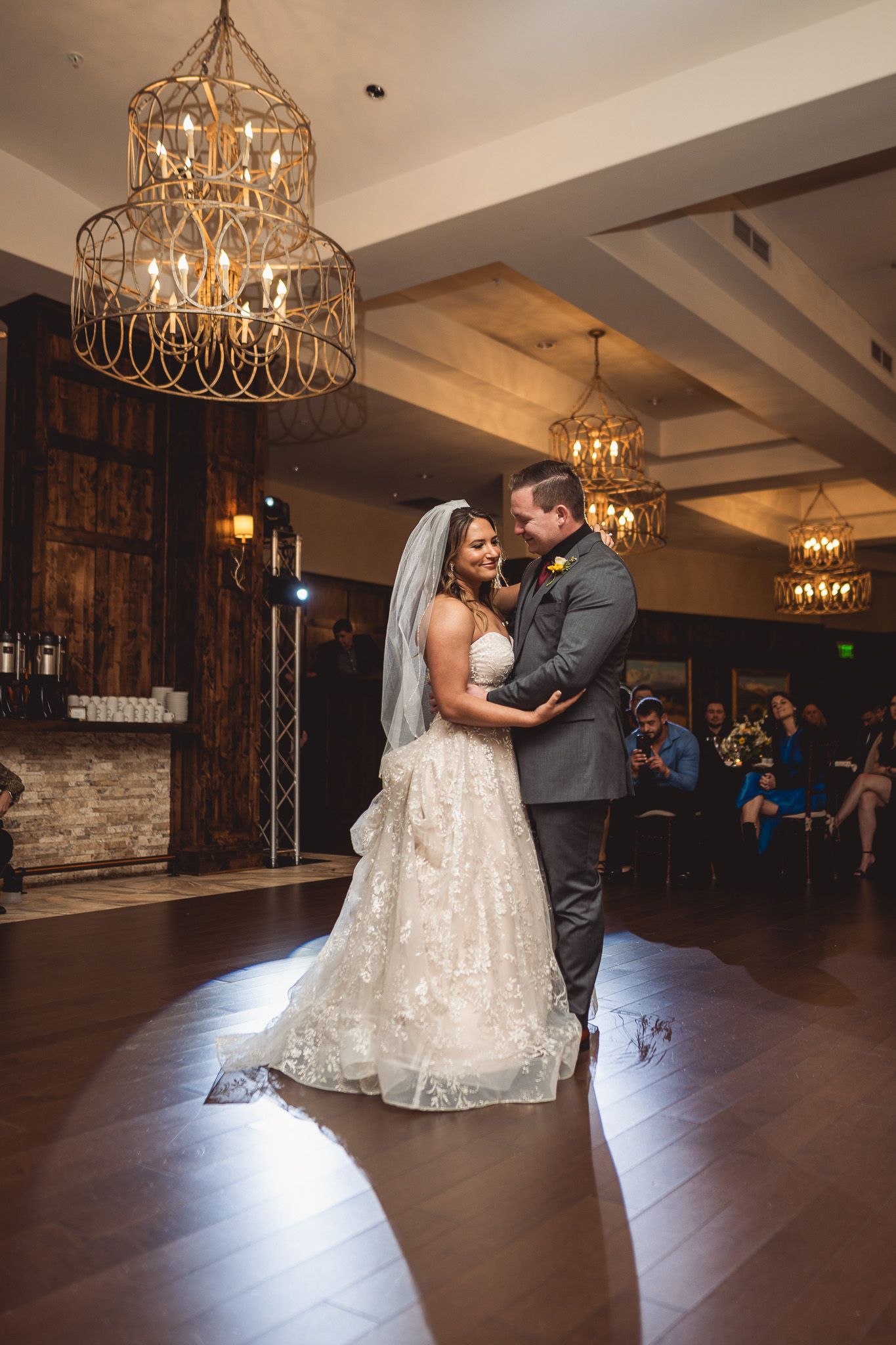 Bride and groom share first dance under elegant gold chandeliers in rustic wedding venue.