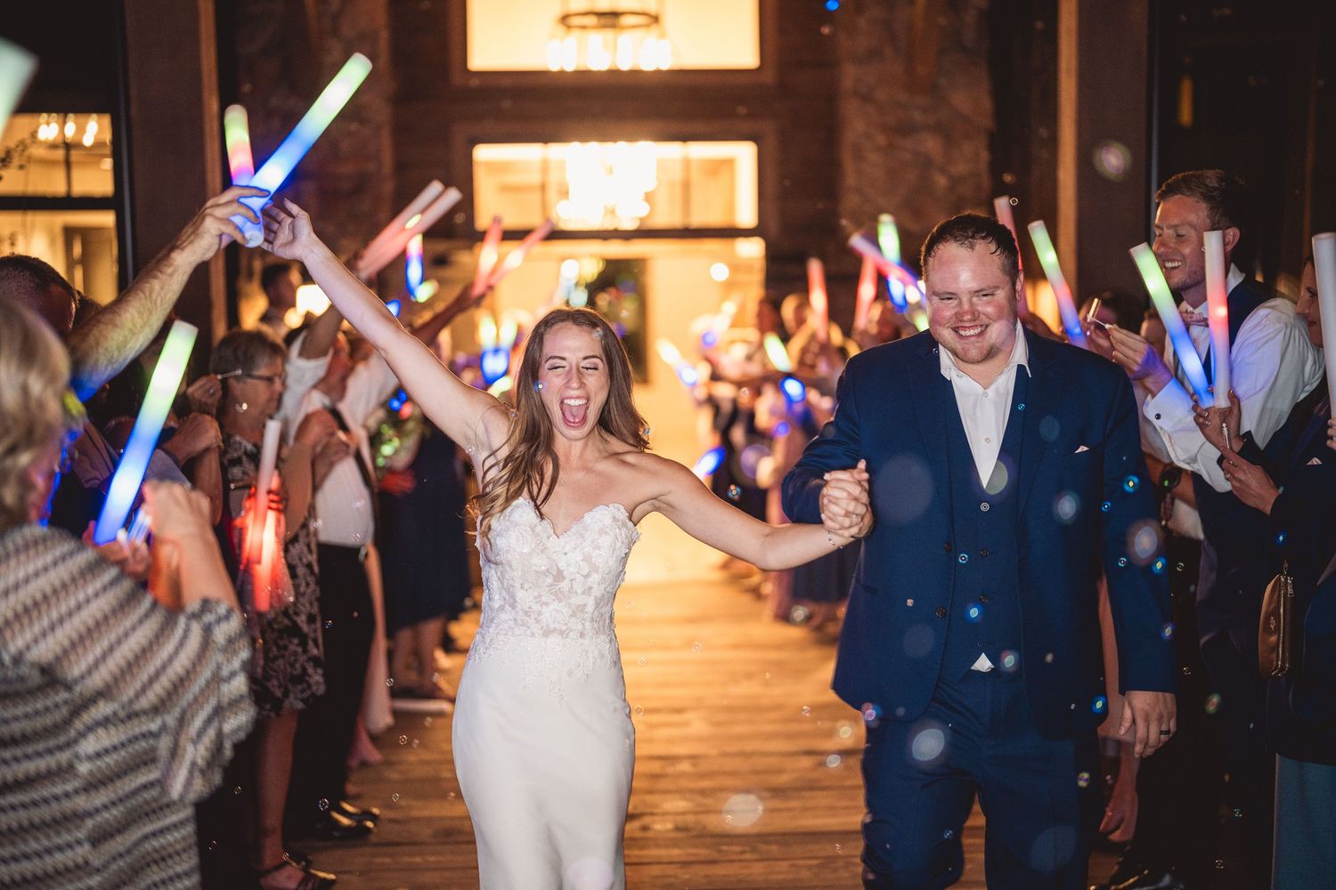 Bride and groom exit wedding reception through a glowing tunnel of colorful light sticks held by guests.